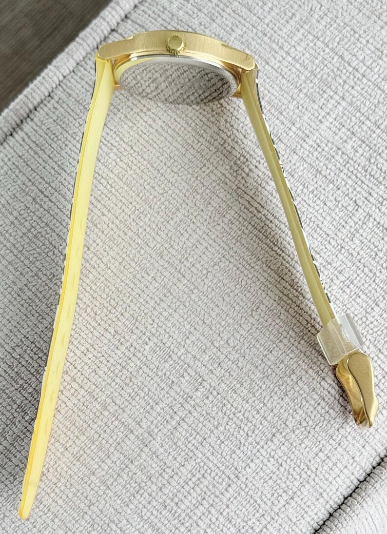 May include: A gold-toned wristwatch with a square face and a yellow patterned band. The watch is lying on a textured surface.