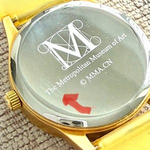 May include: Gold-tone wristwatch with a white face and black lettering. The watch face features a stylized 'M' and the text 'The Metropolitan Museum of Art &copy; MMA.CN'.