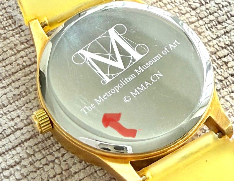 May include: Gold-tone wristwatch with a white face and black lettering. The watch face features a stylized 'M' and the text 'The Metropolitan Museum of Art &copy; MMA.CN'.