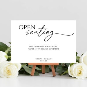 May include: A white sign with black text that reads "OPEN seating" in a cursive font. The sign also includes the text "WE'RE SO HAPPY YOU'RE HERE, PLEASE SIT WHEREVER YOU'D LIKE" and "HANNAH + BENJAMIN 07.03.23". The sign is sitting on a small wooden easel.
