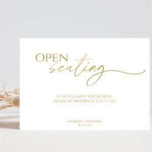 May include: A white sign with gold text that reads "OPEN seating" in a cursive font. The text below reads "WE'RE SO HAPPY YOU'RE HERE PLEASE SIT WHEREVER YOU'D LIKE" and "HANNAH + BENJAMIN 07.03.23".