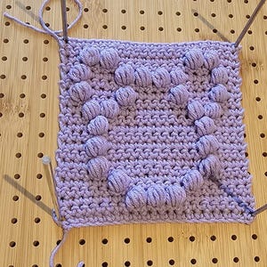 Könnte beinhalten: Ein lila gehäkeltes Quadrat mit einem herzförmigen Muster aus erhabenen Noppen. Das Quadrat wird mit zwei Stricknadeln bearbeitet.