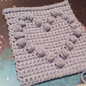 May include: A light blue crocheted square with a textured pattern of raised circles. The square is made with a soft yarn and has a slightly uneven texture.