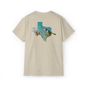 Puede incluir: Una camiseta beige con un estampado gráfico del estado de Texas. El gráfico presenta un pájaro azul volando sobre un cactus con una flor rosa. El texto "CORVINA" está impreso en la esquina superior izquierda del gráfico.