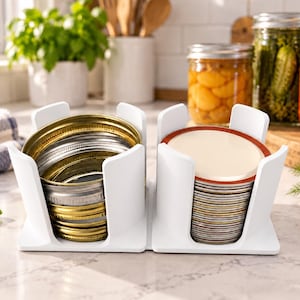 May include: A white plastic organizer with two vertical compartments holding stacks of round metal canning jar lids. The left compartment contains gold-toned rings, while the right compartment holds flat lids with red rubber seals.