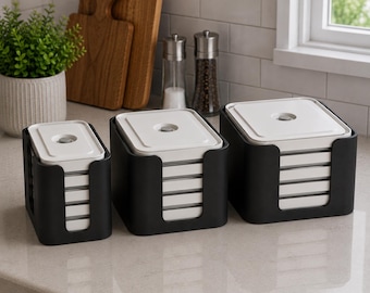 May include: Three square storage containers of varying sizes, each featuring a white lid with a central circular knob, housed within a matte black open-front holder. The containers are stacked in a tiered arrangement on a light-colored countertop.