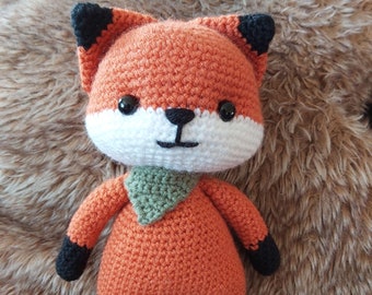May include: A crocheted orange fox plush toy with black eyes, a white muzzle, and a green scarf. The fox is standing on a brown fuzzy surface.