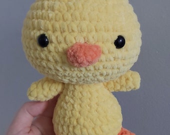 May include: A crocheted yellow chick toy with orange feet and black eyes. The chick is sitting in a hand.