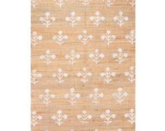 May include: A rectangular woven rug made of natural tan fibers featuring a repeating pattern of white floral motifs arranged in a grid. The texture of the rug is visible, showing the interlaced weave of the material throughout the entire surface.