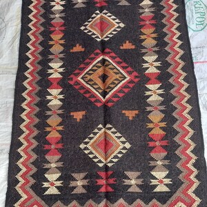 May include: A rectangular, dark brown rug with a geometric pattern. The design features diamond and zig-zag shapes in red, tan, and cream colors. The rug has a fringed edge.