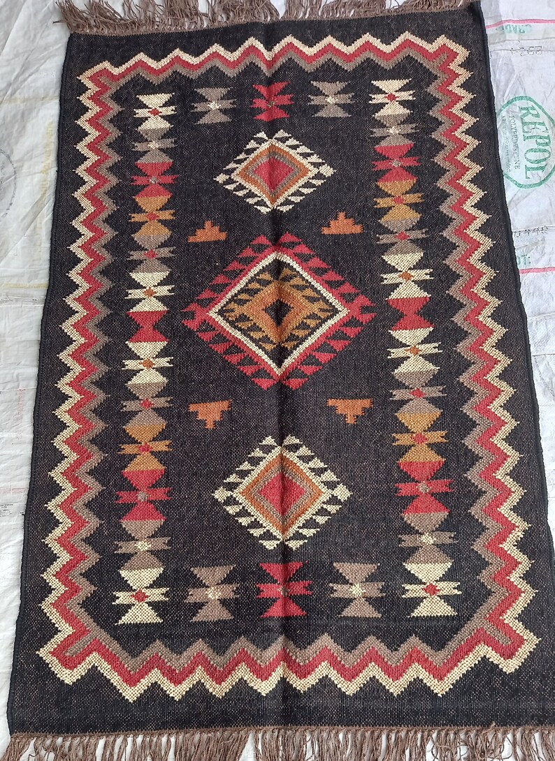 May include: A rectangular, dark brown rug with a geometric pattern. The design features diamond and zig-zag shapes in red, tan, and cream colors. The rug has a fringed edge.