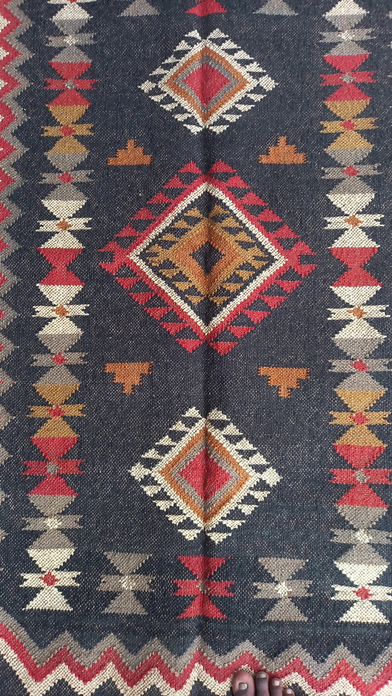May include: A dark blue woven rug with geometric patterns. The rug features diamond and triangle shapes in red, orange, white, and tan. The design is symmetrical, with repeating patterns along the edges.
