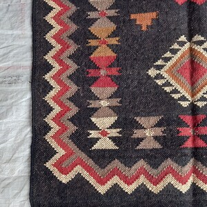 May include: Close-up of a dark gray woven rug with geometric patterns in red, beige, brown, and tan. The rug has a fringed edge and a zig-zag border design. The rug's design features diamond and cross-like shapes.
