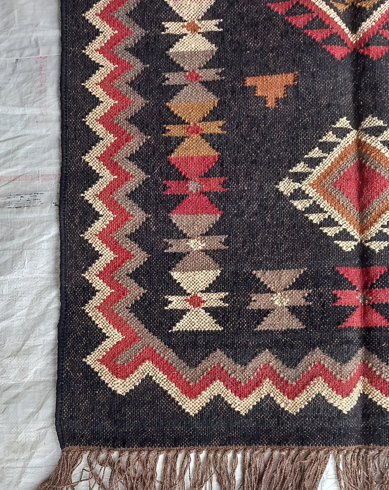 May include: Close-up of a dark gray woven rug with geometric patterns in red, beige, brown, and tan. The rug has a fringed edge and a zig-zag border design. The rug's design features diamond and cross-like shapes.