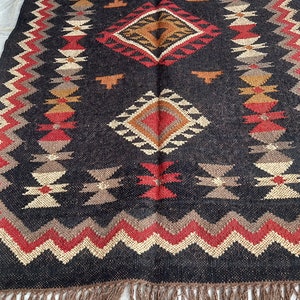 May include: Rectangular black rug with geometric patterns in red, beige, and brown. The rug features diamond and zigzag designs, with a fringe border. The rug is made of woven material, suitable for home decor.
