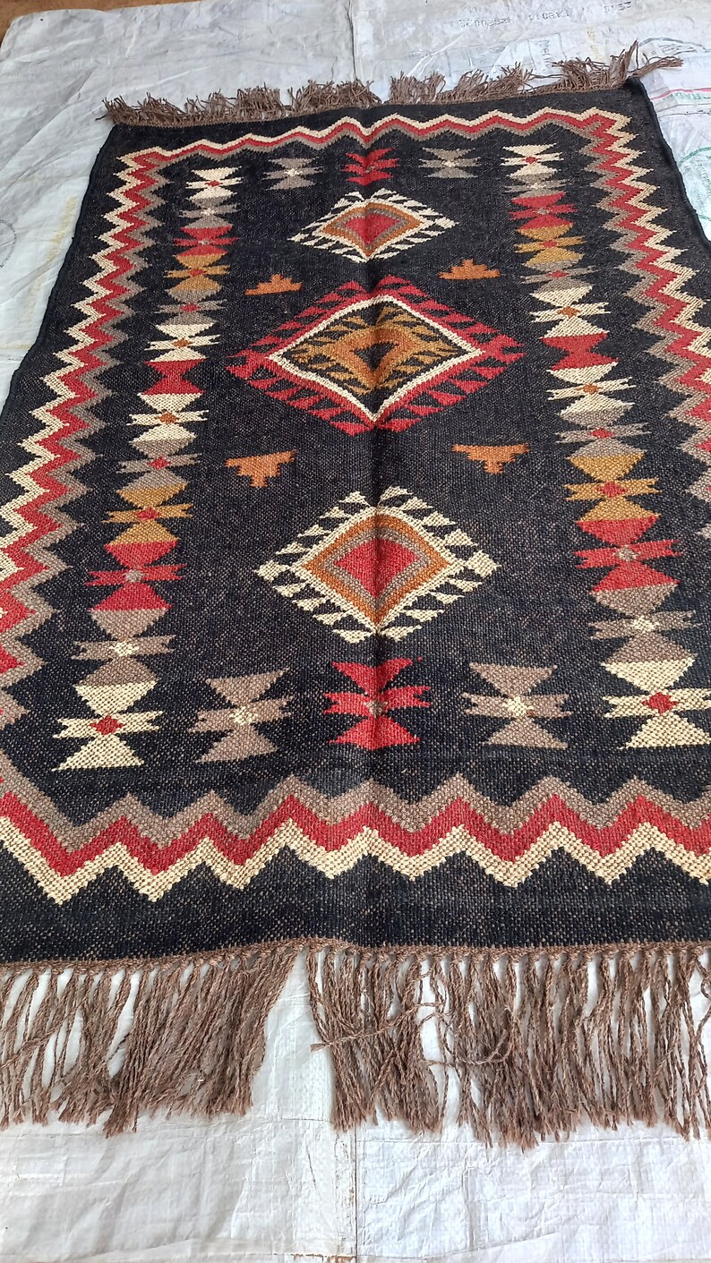 May include: Rectangular black rug with geometric patterns in red, beige, and brown. The rug features diamond and zigzag designs, with a fringe border. The rug is made of woven material, suitable for home decor.