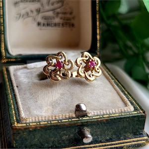 May include: A pair of gold earrings with red gemstone accents in the shape of a flower. The earrings are displayed in a green velvet jewellery box.
