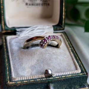 以下が含まれることがあります： 小さな赤い宝石が埋め込まれた、湾曲したバンドのついた金の指輪。指輪は緑色のベルベットの指輪箱に入っています。