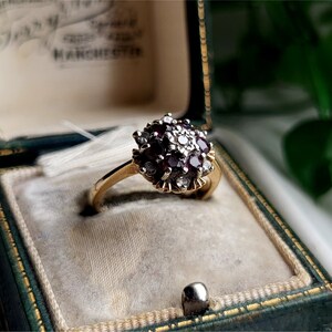 May include: A gold ring with a cluster of red and white gemstones set in a floral design. The ring is in a green velvet ring box.