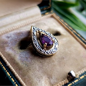 Könnte beinhalten: Ein tropfenförmiger Anhänger mit einem violetten Edelstein, der von einem Heiligenschein aus kleinen, klaren Steinen umgeben ist. Der Anhänger ist in Gold gefasst und wird in einer samtenen Schmuckschatulle präsentiert.