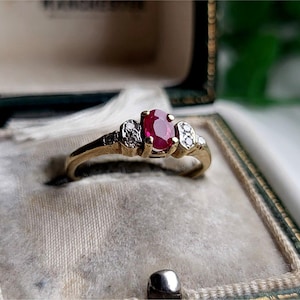 以下が含まれることがあります： 赤い楕円形のルビーと、両側に小さな白いダイヤモンドがついた金の指輪。指輪は緑色のベルベットの指輪箱に入っています。