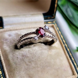 May include: A silver ring featuring a red gemstone and two smaller white gemstones. The ring is displayed in a velvet-lined box.