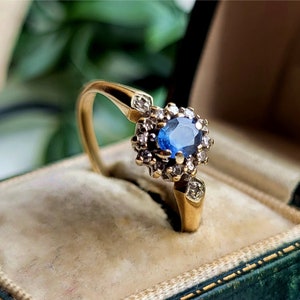 Pode incluir: Um anel de ouro com um safira azul cercado por diamantes menores. O anel est&aacute; em uma caixa de anel de veludo verde.