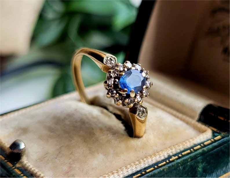 Pode incluir: Um anel de ouro com um safira azul cercado por diamantes menores. O anel est&aacute; em uma caixa de anel de veludo verde.