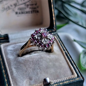 May include: A gold ring with a cluster of red rubies and white diamonds set in a flower shape. The ring is in a green velvet ring box.