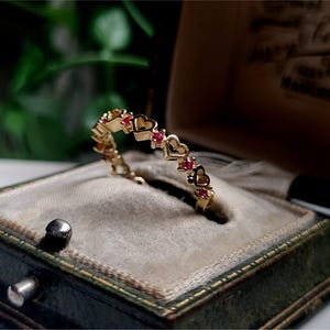 以下が含まれることがあります： 小さな赤い宝石がセットされた、繊細なインターロッキングハートのデザインのゴールドリング。リングは緑色のベルベットのリングボックスに飾られています。