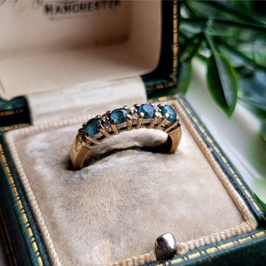 Könnte beinhalten: Ein Goldring mit einer Reihe blauer Edelsteine und kleineren klaren Edelsteinen, die in die Band gefasst sind.