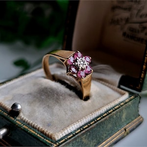 May include: A gold ring with a flower-shaped setting featuring a central diamond surrounded by pink gemstones. The ring is displayed in a green velvet ring box.