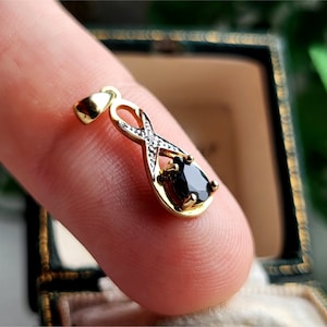 Puede incluir: Colgante de oro con una piedra preciosa negra en forma de lágrima. El colgante tiene un diseño de símbolo de infinito con diamantes. La joya se sostiene entre los dedos, destacando los detalles.