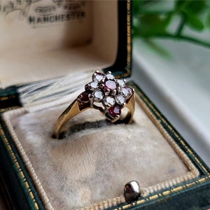 Può includere: Un anello d'oro con un gruppo di pietre preziose rosse e bianche. L'anello è in una scatola per anelli in velluto verde.