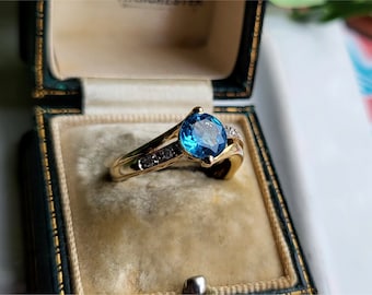 May include: A gold ring with a large blue gemstone surrounded by smaller diamonds. The ring is in a velvet-lined box.