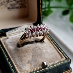 Könnte beinhalten: Ein goldener Ring mit einem Cluster aus roten Rubinen und weißen Diamanten, die in einer Bandfassung gefasst sind. Der Ring befindet sich in einer grünen Samt-Ringbox.