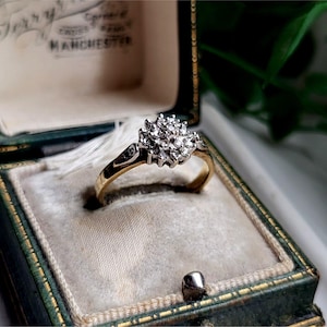 May include: A gold ring with a cluster of small diamonds set in a flower shape. The ring is in a green velvet ring box.