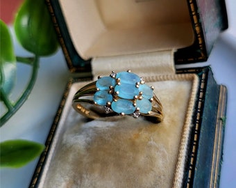May include: A gold ring featuring a cluster of oval-shaped blue gemstones and smaller clear gemstones. The ring is displayed in a velvet ring box.