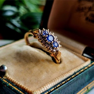 May include: A gold ring featuring a cluster of blue sapphires and diamonds set in a delicate floral design. The ring is presented in a green velvet ring box.
