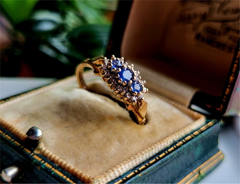 May include: A gold ring featuring a cluster of blue sapphires and diamonds set in a delicate floral design. The ring is presented in a green velvet ring box.