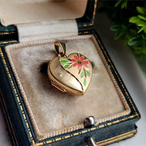 Peut inclure: Un médaillon doré en forme de cœur avec un motif floral rouge, vert et or. Le médaillon est ouvert, révélant un petit espace pour une photo ou un souvenir. Il est présenté dans un écrin à bijoux vintage.