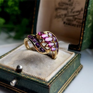 Può includere: Un anello d'oro con un design curvo incastonato con pietre preziose viola e rosa. L'anello è in una scatola per anelli in velluto verde.
