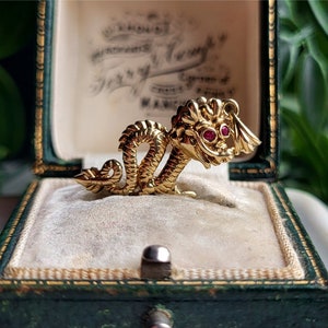 May include: A gold-colored dragon brooch with red gemstone eyes, displayed in a vintage green jewelry box. The dragon is intricately detailed, with a serpentine body and a fierce expression. The box has a cream-colored interior.