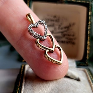 May include: A gold pendant necklace with three interlocking heart charms. The largest heart is plain gold, while the two smaller hearts are accented with small diamonds.