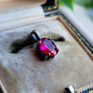 Puede incluir: Un colgante de plata con una vibrante gema roja rubí de corte ovalado. El colgante está engastado en un marco de metal plateado y se exhibe en un joyero vintage con un interior de terciopelo suave. La gema es el punto focal.