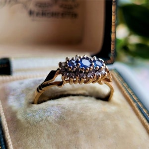 May include: A gold ring featuring three blue sapphires and small diamonds set in a cluster. The ring is displayed in a velvet-lined box.