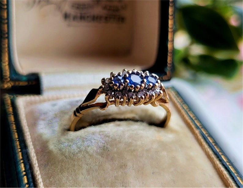 May include: A gold ring featuring three blue sapphires and small diamonds set in a cluster. The ring is displayed in a velvet-lined box.