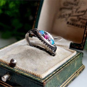 Peut inclure: Une bague en argent avec trois pierres précieuses. La bague a une bande texturée et présente une pierre rose au centre, flanquée de deux opales bleues.