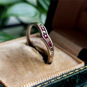 May include: A silver-coloured ring with a curved design, set with small red and clear gemstones. The ring is displayed in a velvet-lined jewellery box.