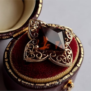 May include: A gold filigree pendant with a large, dark brown gemstone in the center. The pendant is in a red velvet jewelry box.